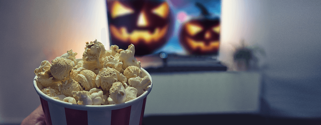 Popcorn in einem rot-weiß-gestreiften Becher wird im Vordergrund von einer Hand gehalten, während im Hintergrund ein Halloween-Kürbis im Fernsehen auf einem weißen Sideboard zu sehen ist.